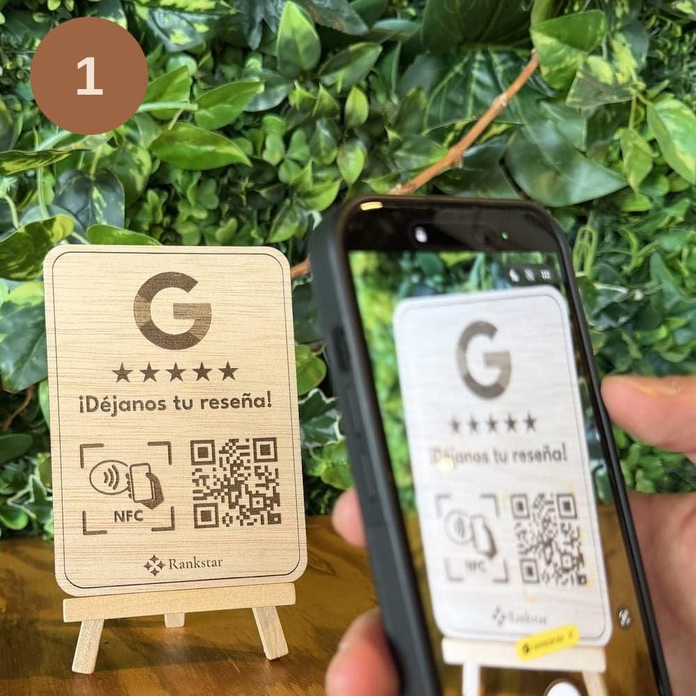 Una mano escanea con el móvil el QR del expositor de madera de Rankstar con el logo de Google y «¡Déjanos tu reseña!», sobre una mesa delante de una pared de plantas