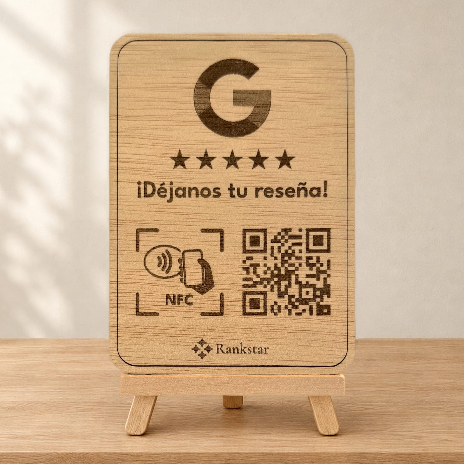 Expositor Rankstar de madera sobre su caballete, con el logo de Google, cinco estrellas, el rótulo «¡Déjanos tu reseña!», el símbolo NFC y el QR grabados a láser, sobre una mesa de madera clara junto a una pared con sombras de hojas
