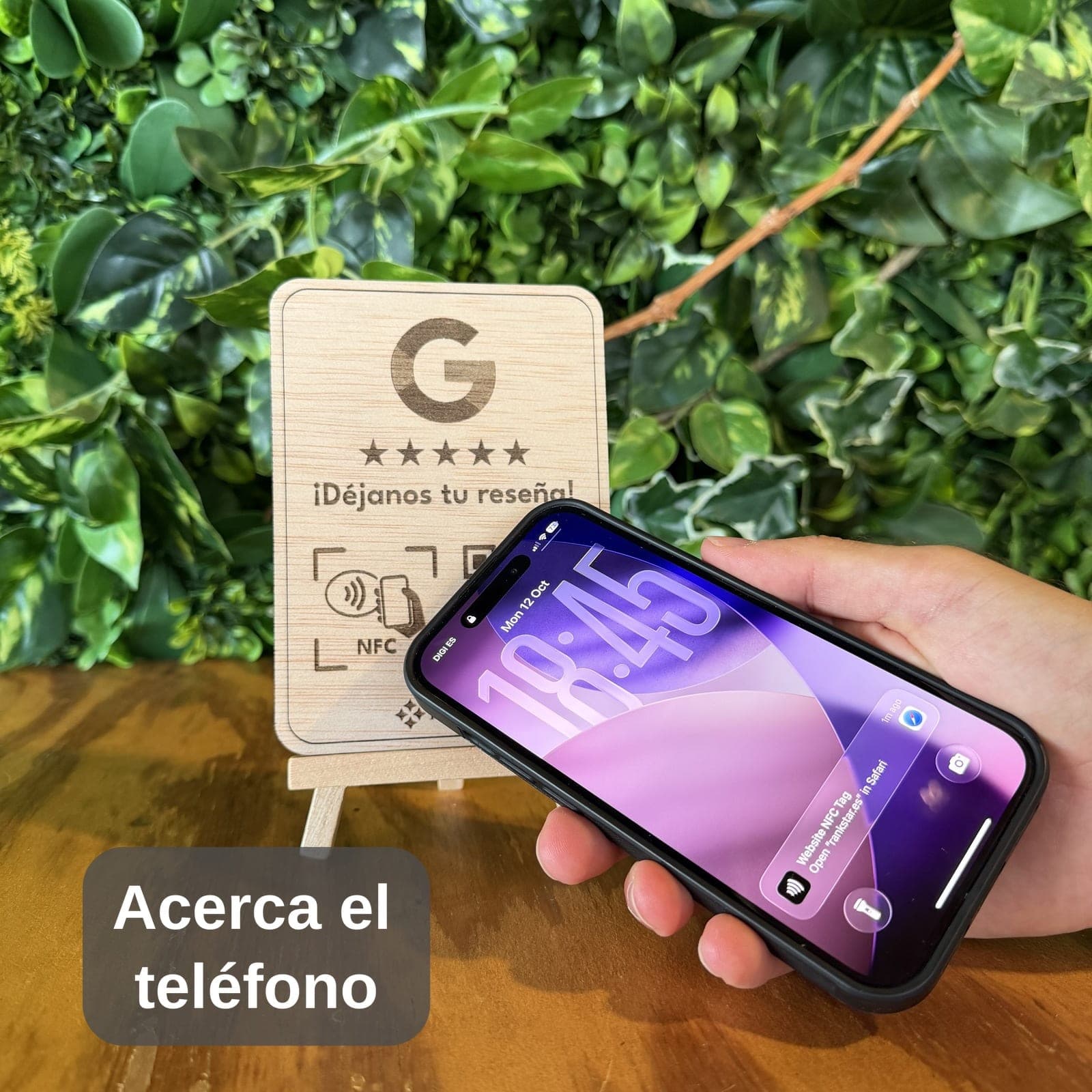 Una mano acerca el móvil al expositor Rankstar de Google sobre una mesa, delante de una pared de plantas, con el rótulo «Acerca el teléfono»