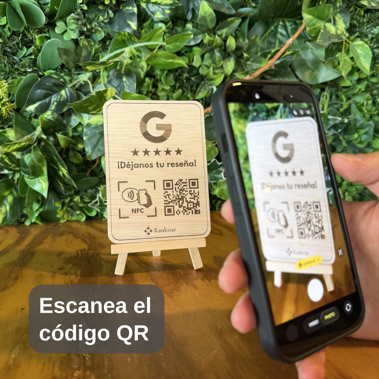 Una mano escanea con el móvil el QR del expositor Rankstar de Google sobre una mesa, delante de una pared de plantas, con el rótulo «Escanea el código QR»