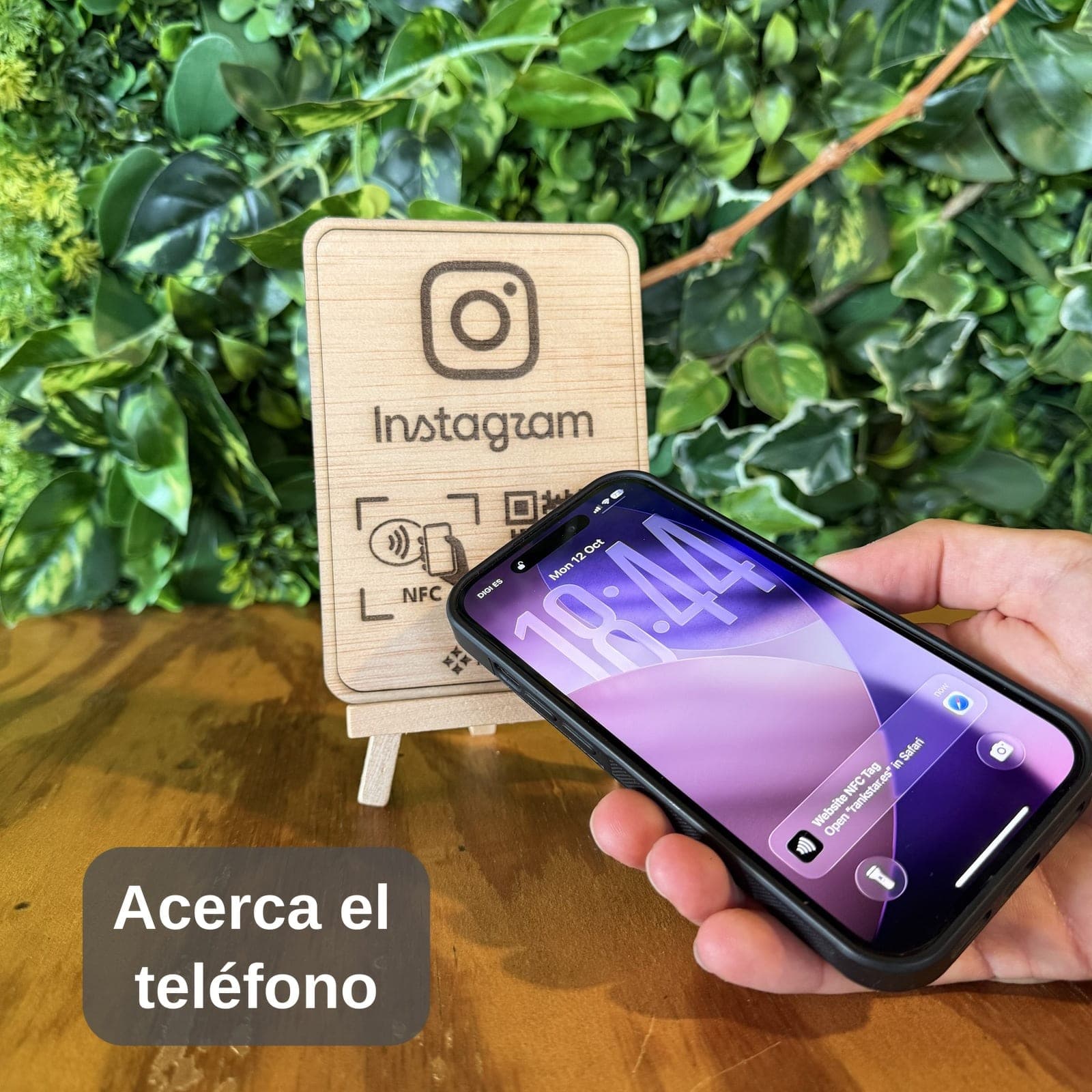 Una mano acerca el móvil al expositor Rankstar de Instagram sobre una mesa, delante de una pared de plantas, con el rótulo «Acerca el teléfono»