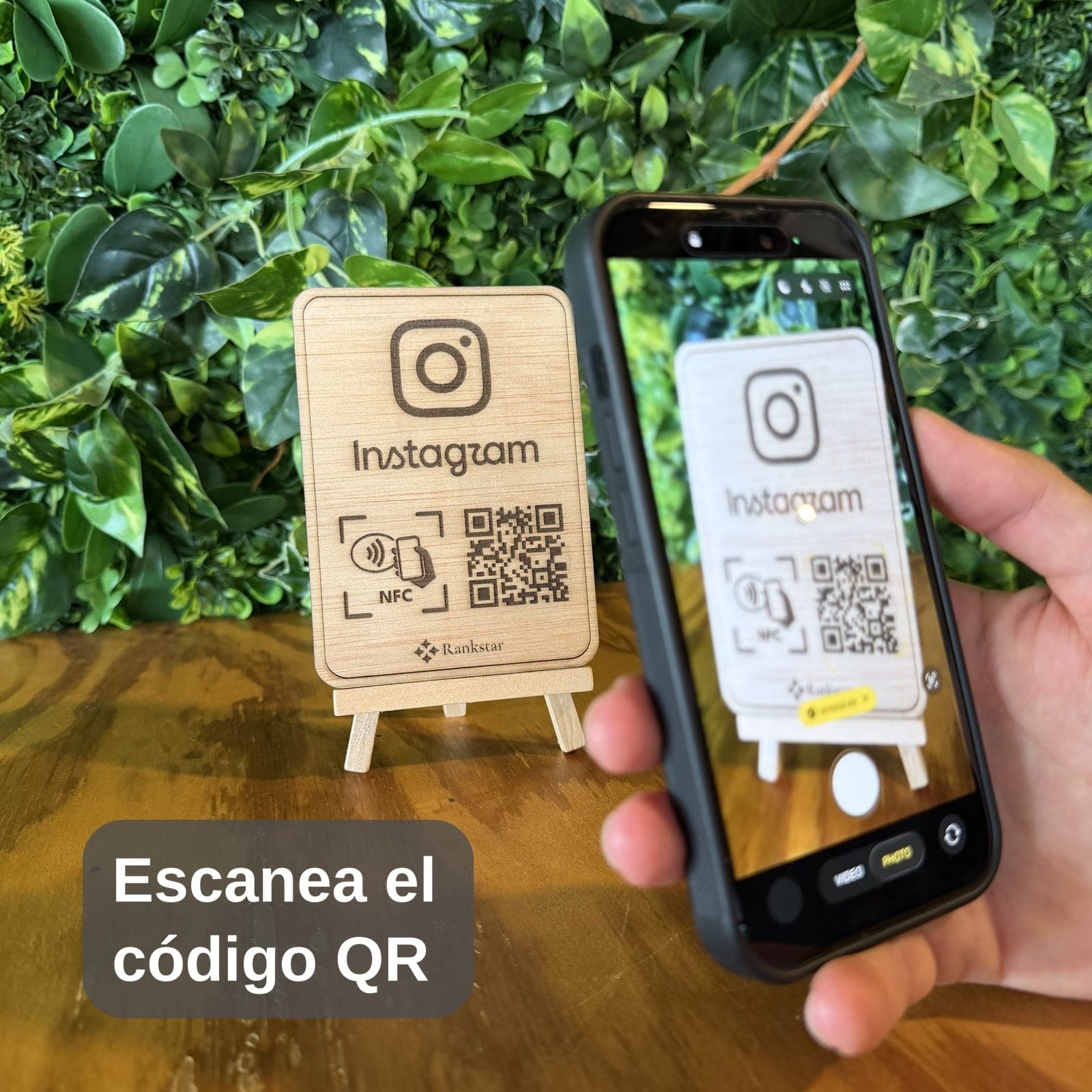 Una mano escanea con el móvil el QR del expositor Rankstar de Instagram sobre una mesa, delante de una pared de plantas, con el rótulo «Escanea el código QR»
