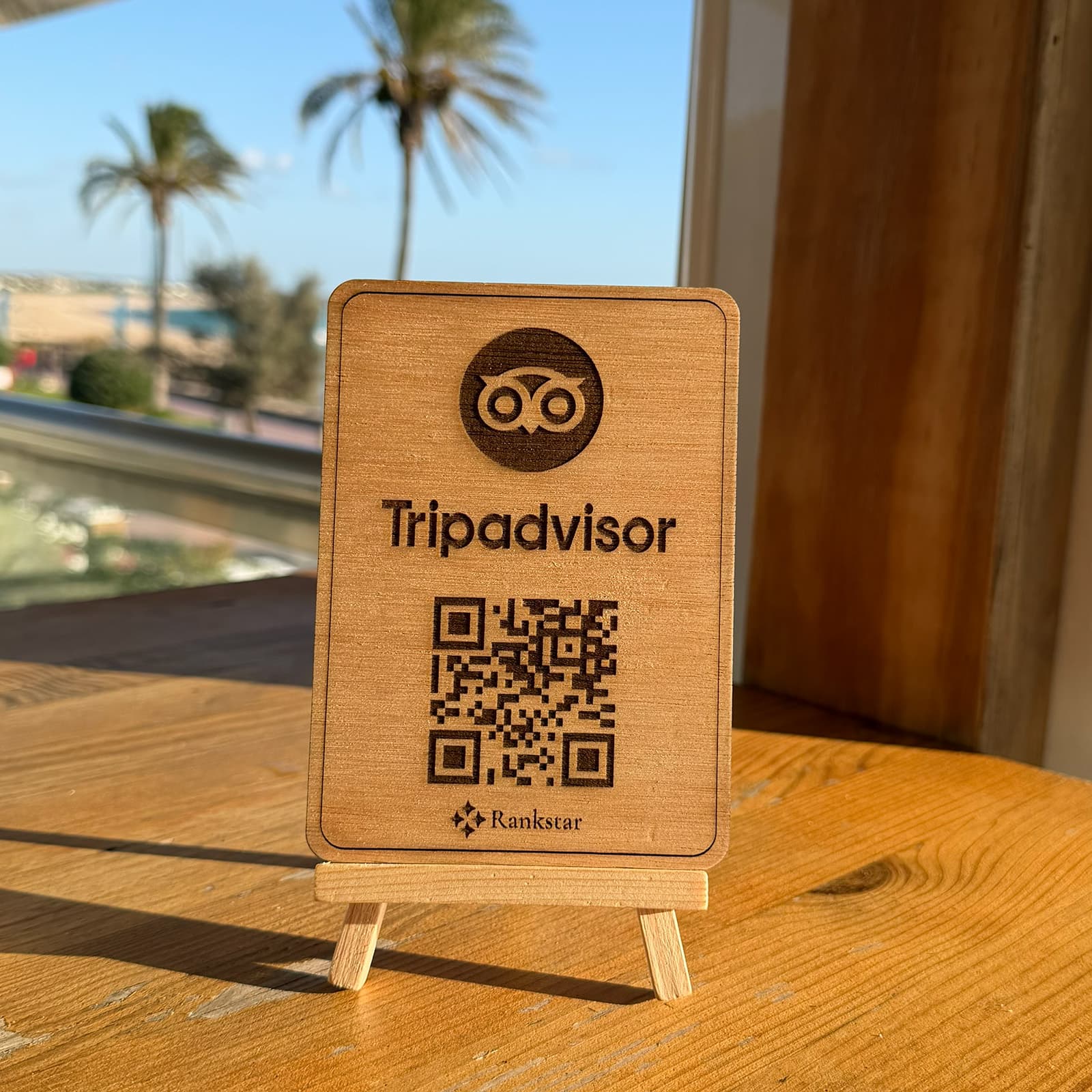 Expositor Rankstar de Tripadvisor sobre la mesa de una terraza, con palmeras y el mar al fondo