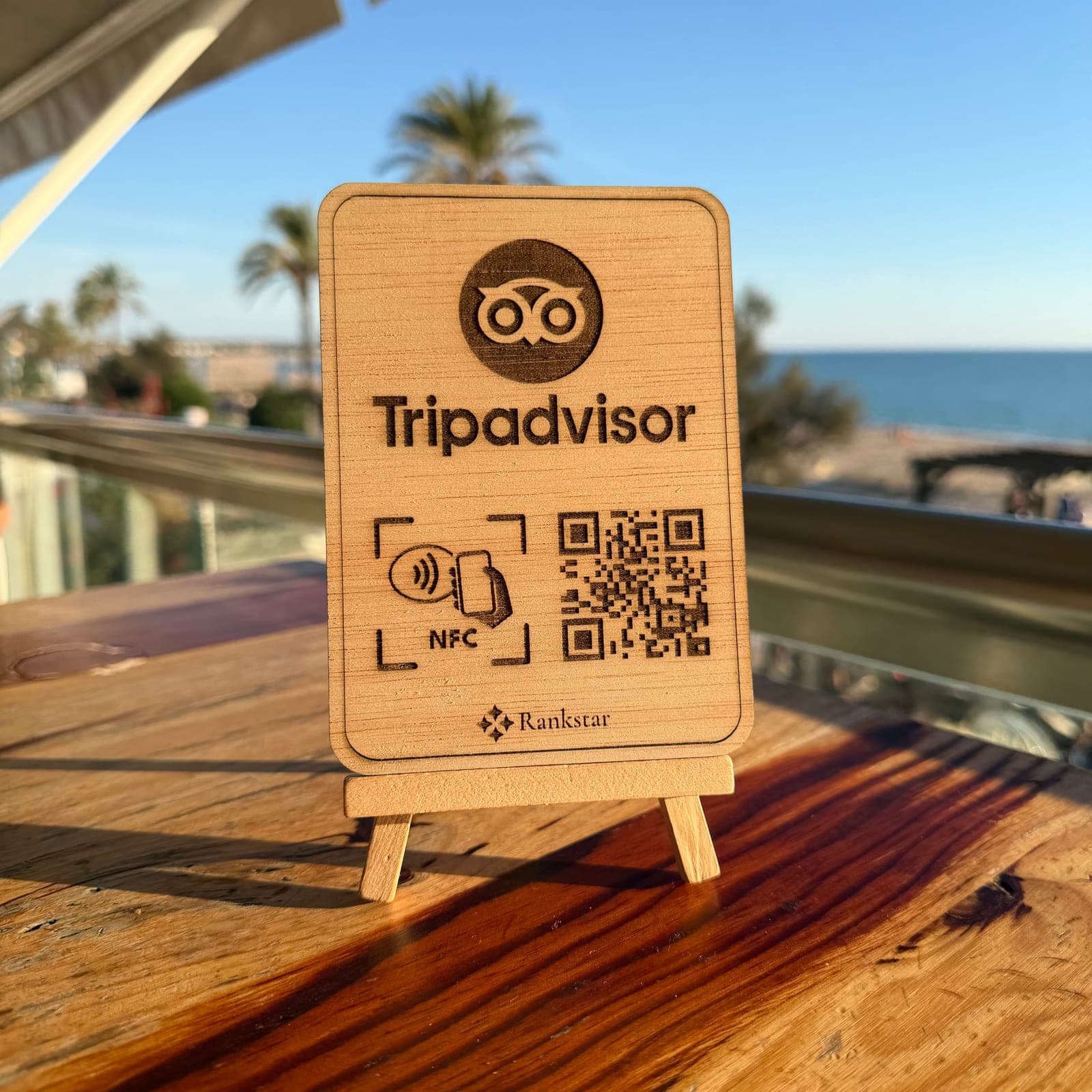 Expositor Rankstar de Tripadvisor en la mesa de una terraza, con palmeras y el mar al fondo