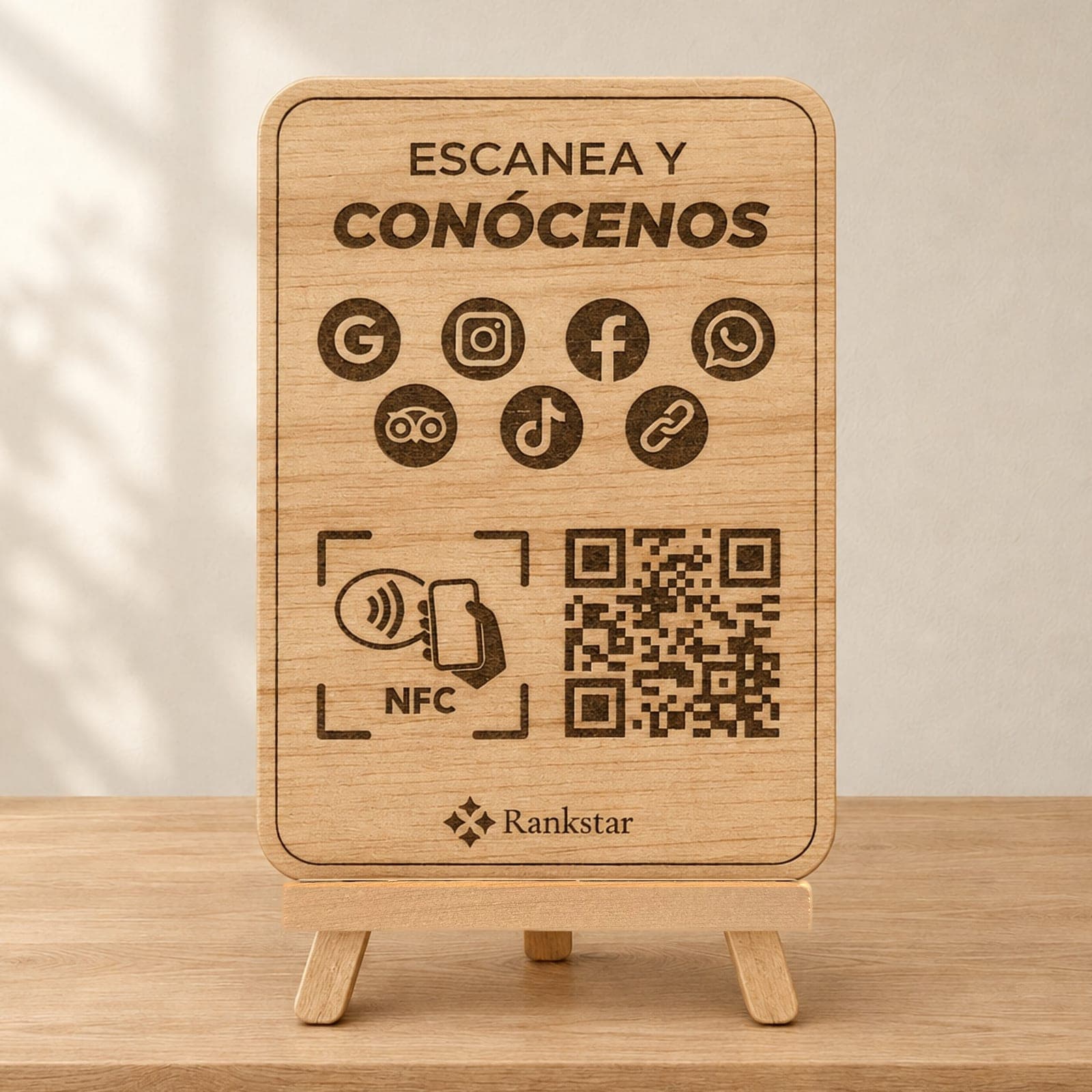 Expositor Rankstar de madera sobre su caballete, con el rótulo «Escanea y conócenos», los iconos de Google, Instagram, Facebook, WhatsApp, Tripadvisor y TikTok, el símbolo NFC y el QR grabados a láser, sobre una mesa de madera clara junto a una pared con sombras de hojas