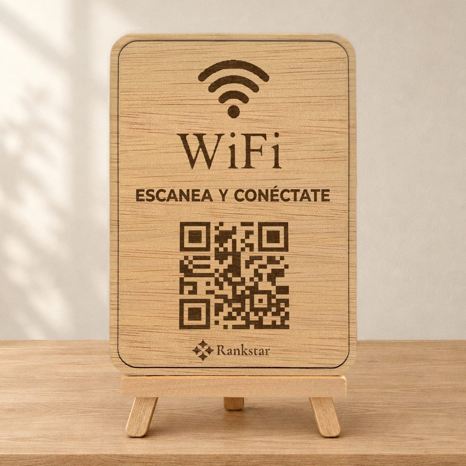 Expositor Rankstar de madera sobre su caballete, con el icono de WiFi, el rótulo «Escanea y conéctate» y el QR grabados a láser, sobre una mesa de madera clara junto a una pared con sombras de hojas