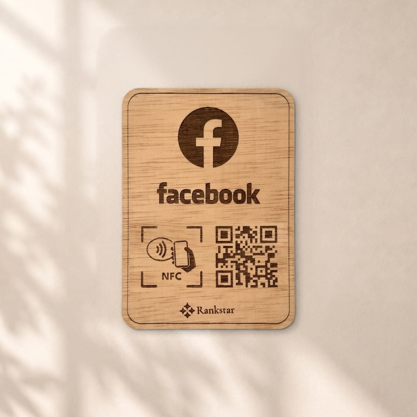 Placa Rankstar de madera con el logo de Facebook, el símbolo NFC y el QR grabados a láser, en una pared clara con sombras de hojas