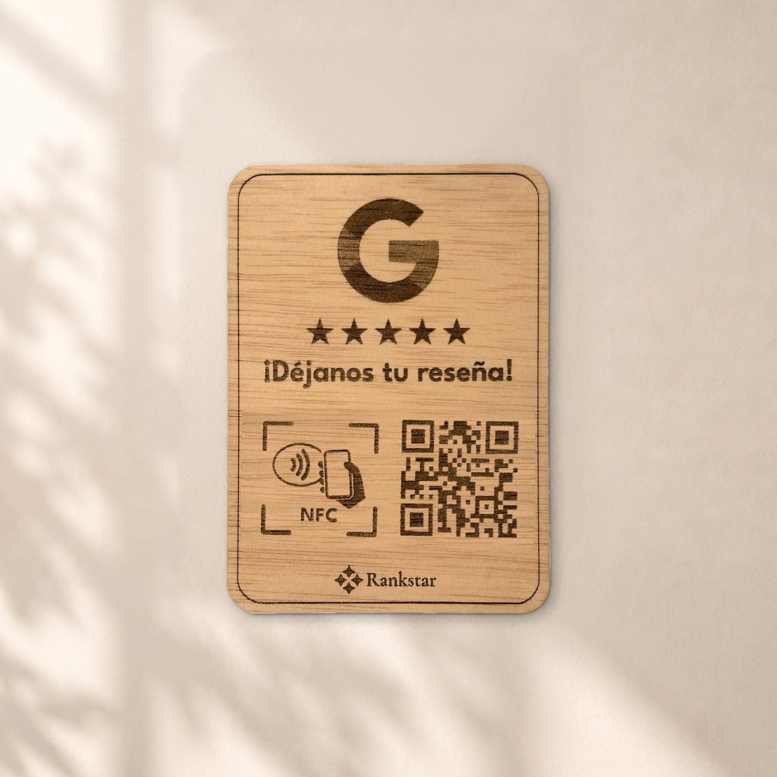 Placa Rankstar de madera con el logo de Google, cinco estrellas, el rótulo «¡Déjanos tu reseña!», el símbolo NFC y el QR grabados a láser, en una pared clara con sombras de hojas