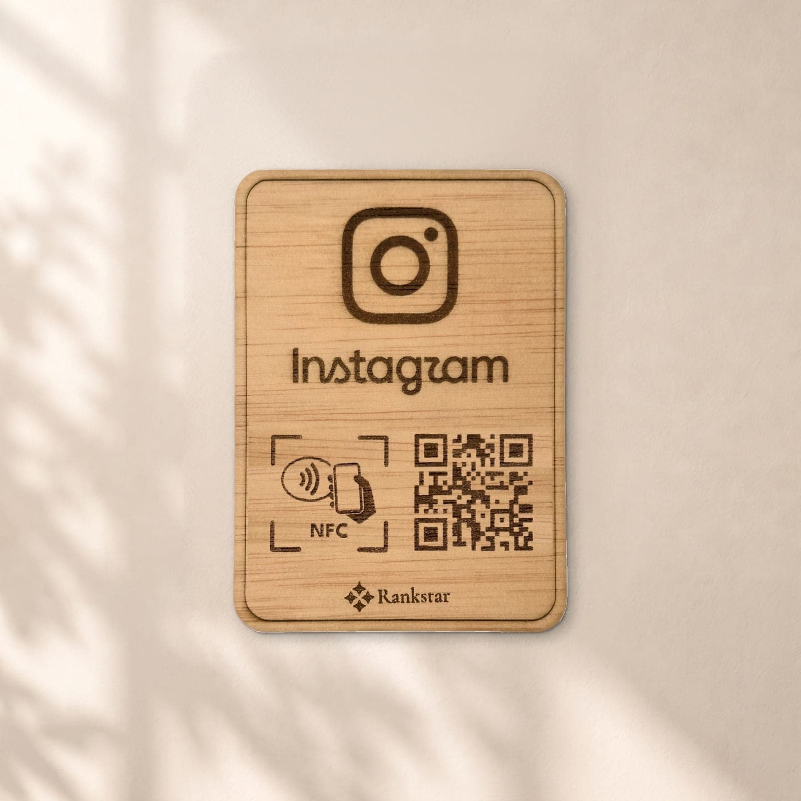 Placa Rankstar de madera con el logo de Instagram, el símbolo NFC y el QR grabados a láser, en una pared clara con sombras de hojas