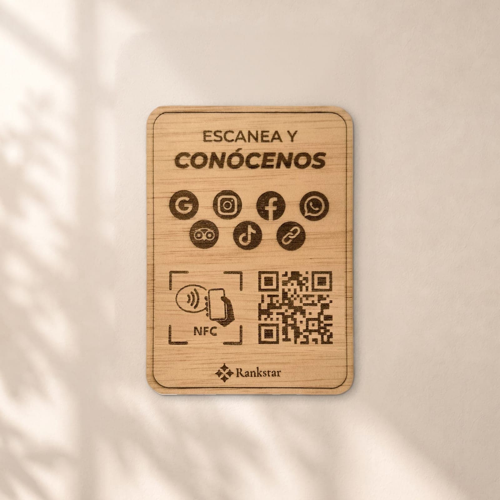 Placa Rankstar de madera con el rótulo «Escanea y conócenos», los iconos de Google, Instagram, Facebook, WhatsApp, Tripadvisor y TikTok, el símbolo NFC y el QR grabados a láser, en una pared clara con sombras de hojas