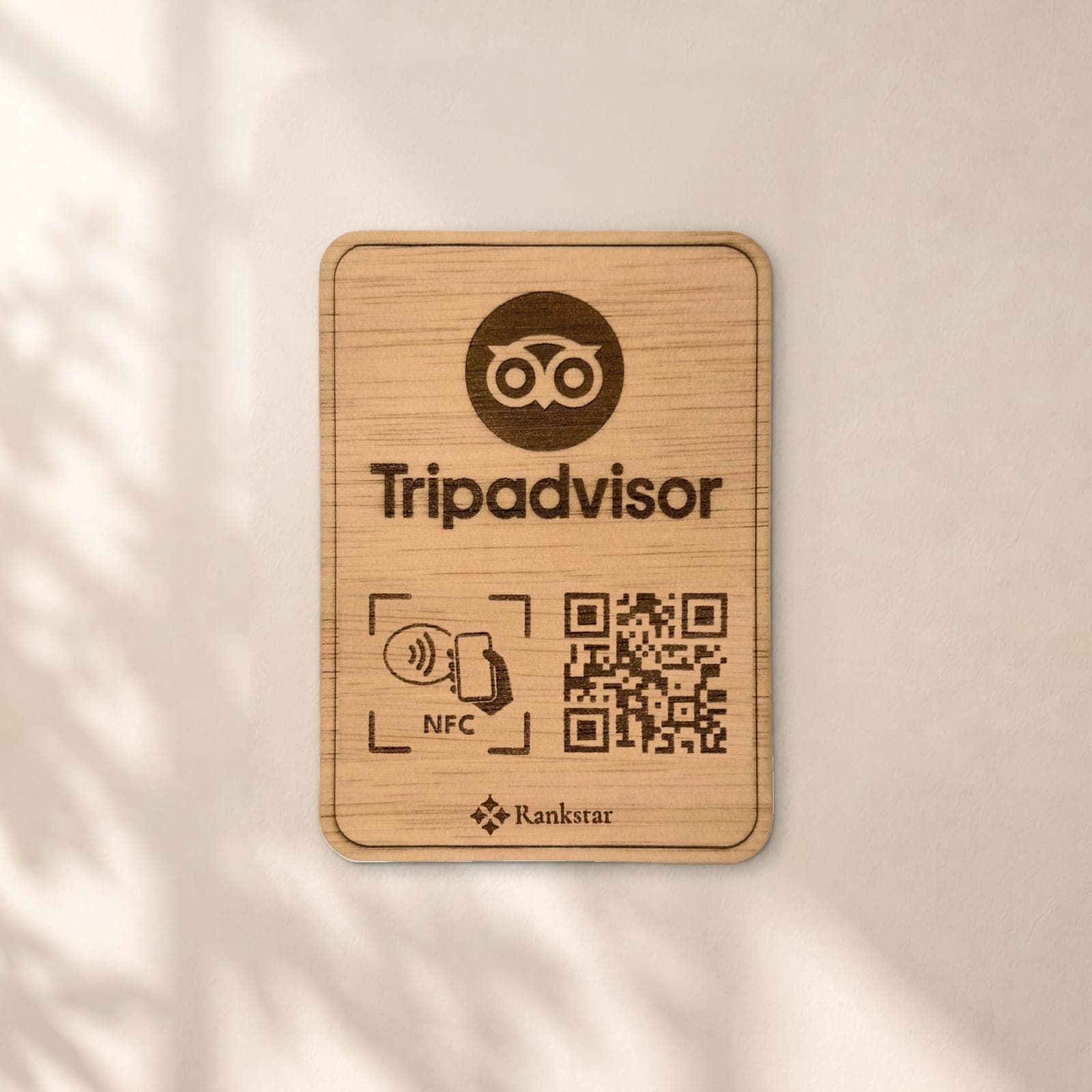 Placa Rankstar de madera con el logo de Tripadvisor, el símbolo NFC y el QR grabados a láser, en una pared clara con sombras de hojas
