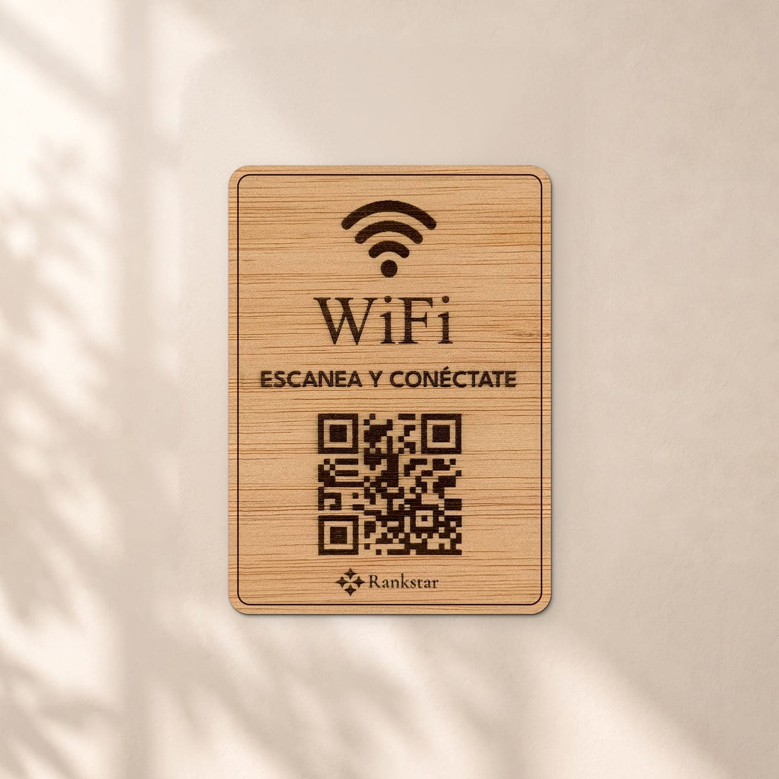 Placa Rankstar de madera con el icono de WiFi, el rótulo «Escanea y conéctate» y el QR grabados a láser, en una pared clara con sombras de hojas